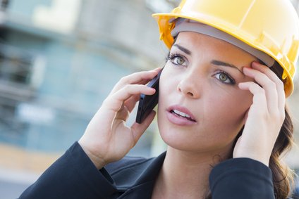 Young Worried Looking Professional Female Contractor Wearing Hard Hat at Construction Site Using Cell Phone.